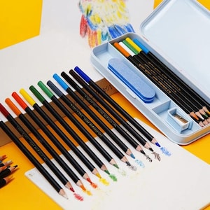 Puede incluir: Un juego de lápices de colores con barriles negros, dispuestos sobre una superficie blanca. Los lápices son de varios colores, incluyendo rojo, naranja, amarillo, verde y azul. Un estuche azul contiene lápices adicionales, una goma de borrar y un sacapuntas. Los lápices son para dibujar y arte.