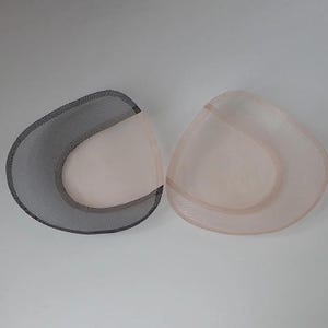 May include: Two triangular bra cup inserts, one featuring a black mesh outer layer and the other a beige mesh outer layer. Both inserts have a smooth, solid-coloured interior lining and a curved, moulded shape designed for garment support.