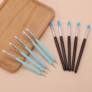 May include: A set of ten sculpting tools featuring five brushes with light blue handles and white silicone tips, and five brushes with dark brown handles and light blue silicone tips. Each tool has a silver metal ferrule connecting the tip to the handle.