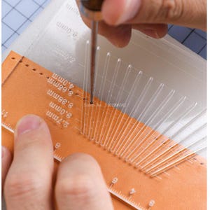 Peut inclure: Gros plan sur un processus de fabrication de cuir. Une règle en plastique transparent avec des marquages de mesure est placée sur du cuir orange. Une main tient un outil, perçant le cuir. La règle a des lignes d'espacement.
