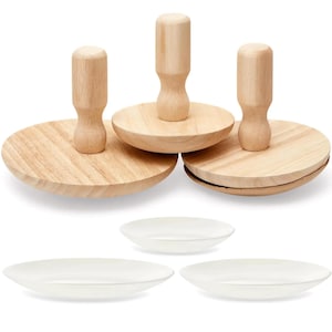 May include: A set of three wooden food presses with rounded handles and circular bases, alongside three white, oval-shaped serving plates. The presses are made of light-colored wood, and the plates are smooth and glossy.