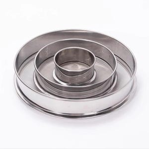 May include: A set of three circular, silver-coloured metal sieves nested inside one another. Each sieve features a smooth, polished rim and a fine mesh screen base. The metallic surfaces reflect light, showing a clean, industrial design.