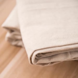 Puede incluir: Una pieza doblada de tela cruda de color blanquecino con una textura tejida visible. El material está superpuesto, mostrando múltiples pliegues y arrugas suaves a lo largo de los bordes, con un color beige claro natural en toda la superficie.
