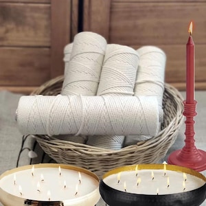 May include: A woven wicker basket containing several spools of white cotton macrame cord, accompanied by two large round candles with multiple lit wicks and a single tall red taper candle standing in a matching red candle holder.