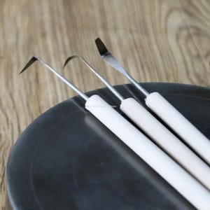 May include: Three metal sculpting tools with white cylindrical handles. Each tool features a distinct stainless steel tip: one pointed and angled, one curved and hooked, and one flat and spatula-shaped, all arranged side by side.