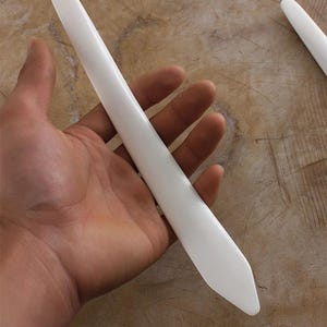 May include: A white bone folder tool, held in a hand. The tool is smooth and tapered, with a rounded handle and a pointed tip. The background is a light brown surface.