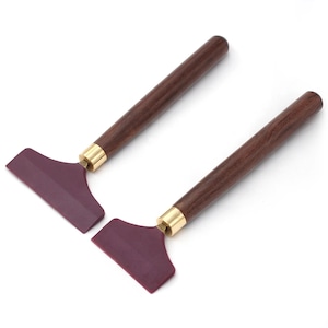 May include: Two artist palette knives with dark wood handles, brass ferrules, and burgundy blades. The tools are angled slightly, showcasing their design and craftsmanship. The background is plain white.