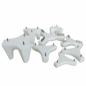 May include: A collection of white, three-legged ceramic kiln stilts with pointed metal pins protruding from the top surfaces. These tripod-shaped supports vary in size and are arranged in a cluster, featuring smooth, matte white surfaces.