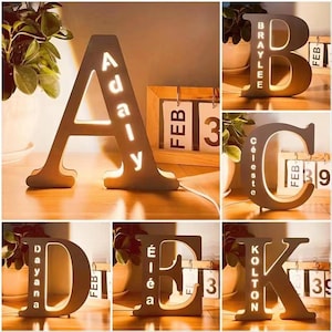 Pode incluir: Lâmpadas de letras de madeira em forma das letras A, B, C, D, E e K. Cada letra tem um nome recortado e iluminado com um brilho quente. Um calendário de madeira está no fundo.
