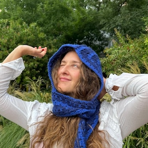 Peut inclure: Ensemble bonnet et écharpe crochet bleu. Le bonnet présente un motif à carreaux dans les tons de bleu. L'écharpe est longue et s'enroule autour du cou. L'ensemble est en fil doux et texturé. La personne porte un chemisier blanc.