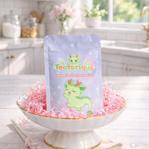 May include: A light purple tea packet with the text "Teafarious" and a cartoon dragon illustration. The packet is displayed in a white and gold footed bowl filled with pink paper shreds. The words "Instant Potion Packed Tea" are also on the packet.