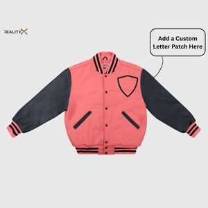 May include: A coral-colored varsity jacket with dark gray sleeves. The jacket features a black shield-shaped patch, black trim, and the text "Add a Custom Letter Patch Here". The jacket has a button-up front and striped cuffs.