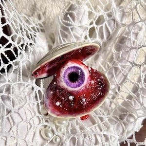 May include: An open seashell containing a realistic human-like eye with a vibrant purple iris and black pupil. The interior of the shell is textured with deep red tones and scattered small white pearls, resting against a white lace fabric background.