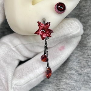 May include: A set of three earrings featuring a dark red flower stud, a red eye-shaped stud, and a dangling earring with a red flower charm attached to two thin dark metal chains ending in small, faceted red teardrop-shaped crystals.