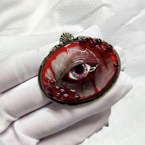 Könnte beinhalten: Eine ovale Brosche mit einem realistischen menschlichen Auge mit blauer Iris, eingebettet in einen glänzenden, blutroten und weißen Harzhintergrund. Das Stück ist in einem verzierten, antik wirkenden Bronzerahmen mit kleinen, erhabenen roten Tropfen gefasst.