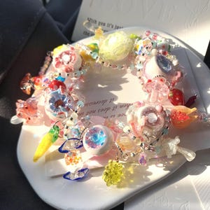 May include: A colorful beaded bracelet with various charms, including flowers, fruits, and abstract shapes. The bracelet features a mix of pink, blue, yellow, and clear beads. A card with the word "LOVE" and text is in the background.