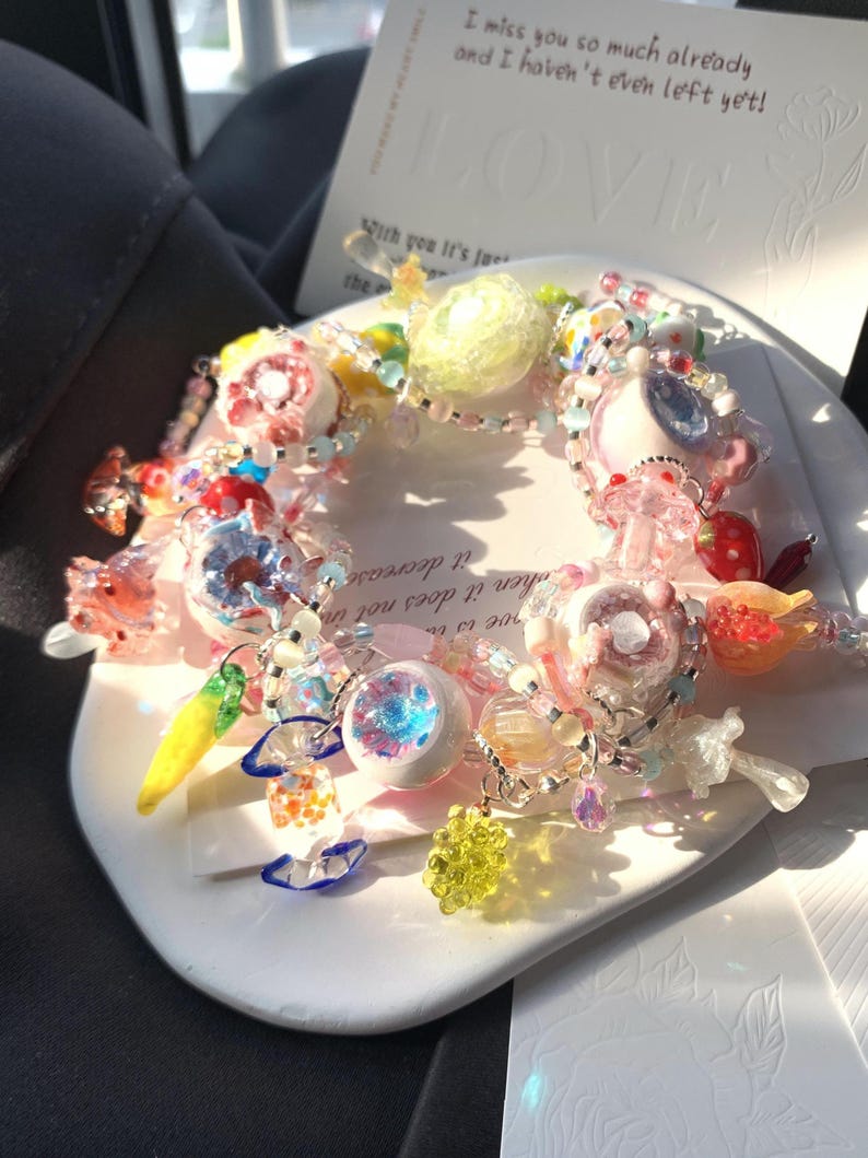 May include: A colorful beaded bracelet with various charms, including flowers, fruits, and abstract shapes. The bracelet features a mix of pink, blue, yellow, and clear beads. A card with the word "LOVE" and text is in the background.