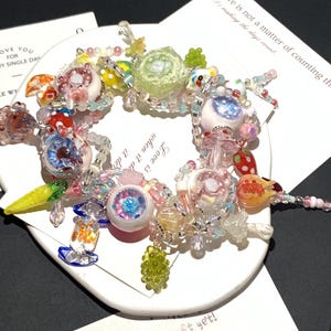 May include: A colorful beaded bracelet with various charms, including eye-shaped beads, a green rose, and fruit-shaped beads. The bracelet is on a white circular surface with text that reads "Love is not a matter of counting the days count."