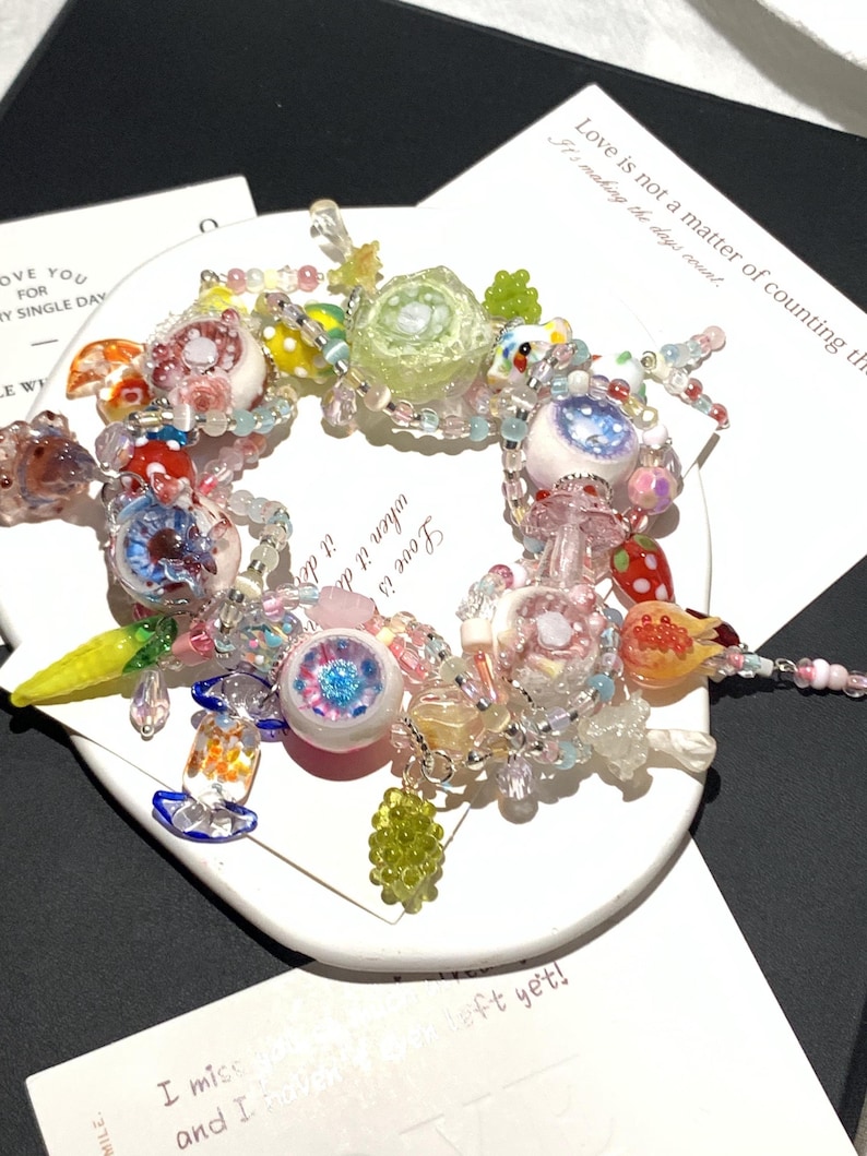 May include: A colorful beaded bracelet with various charms, including eye-shaped beads, a green rose, and fruit-shaped beads. The bracelet is on a white circular surface with text that reads "Love is not a matter of counting the days count."