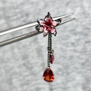 May include: A dark metal ear cuff featuring a white and red enameled flower accent. Two delicate dark metal chains hang from the flower, each ending in a faceted, teardrop-shaped red gemstone. The piece is held by silver-toned metal tweezers.