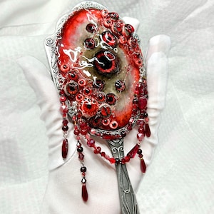May include: A decorative hand mirror with a silver handle and frame. The mirror's surface is adorned with red and black elements, including multiple red eyes and beaded accents. The handle has an ornate design.