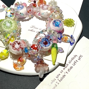 May include: A colorful beaded bracelet with various glass charms, including eye-shaped beads, mushrooms, and fruit shapes. The bracelet is on a white surface with handwritten notes in the background. The bracelet features a mix of pink, blue, yellow, and clear beads.