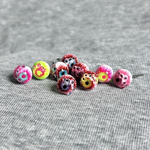 May include: A collection of colorful eyeball-shaped beads. The beads feature various designs, including pink, yellow, and red with intricate details. Some have blue, yellow, and red irises. The beads are arranged on a gray textured surface.