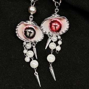 May include: A pair of silver-toned hoop earrings featuring white seashell charms. Each shell contains a realistic eyeball with a cross-shaped pupil, surrounded by dangling white faux pearls and small silver-toned metal spikes.