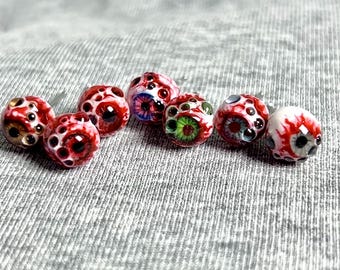 May include: A collection of handmade glass beads, each designed to resemble a human eye. The beads feature intricate details with red, white, and black color schemes, with some beads showcasing blue and green irises. These unique beads are ideal for jewelry making.