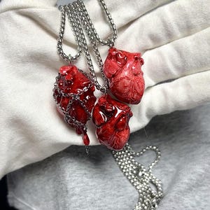 May include: Three red heart-shaped pendants on silver chains. One heart is encased in a silver chain, another has a dripping effect, and the third is a detailed anatomical model. The pendants are displayed against a white and gray background.