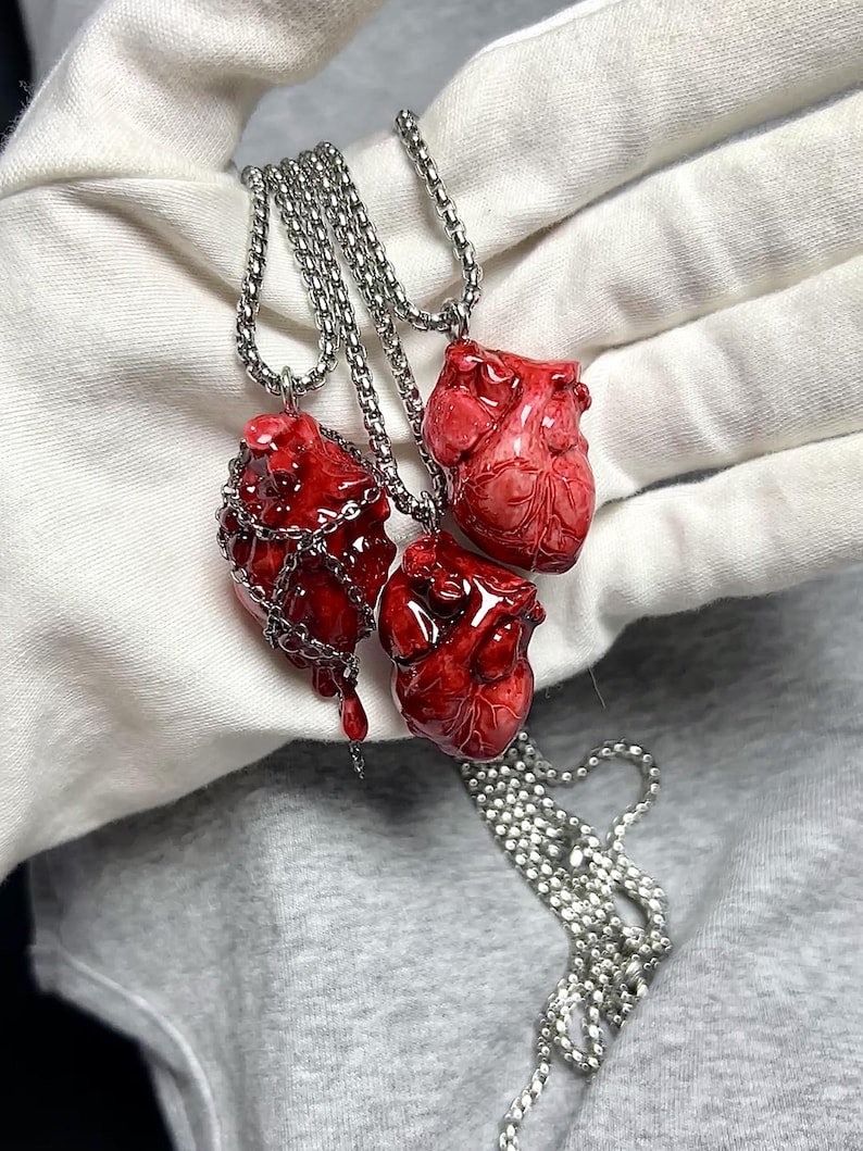 May include: Three red heart-shaped pendants on silver chains. One heart is encased in a silver chain, another has a dripping effect, and the third is a detailed anatomical model. The pendants are displayed against a white and gray background.