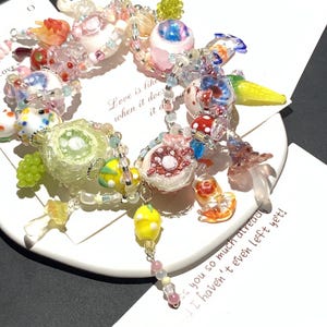 May include: A colorful beaded bracelet with various charms, including fruit, mushrooms, and abstract shapes. The bracelet features a mix of clear, pink, yellow, and blue beads. Text on a card reads "Love is like when it does it does".