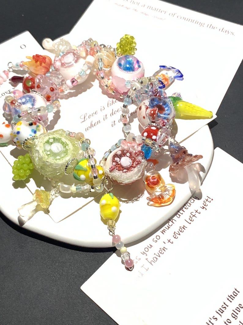 May include: A colorful beaded bracelet with various charms, including fruit, mushrooms, and abstract shapes. The bracelet features a mix of clear, pink, yellow, and blue beads. Text on a card reads "Love is like when it does it does".
