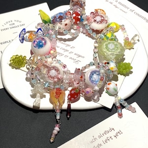 May include: A colorful beaded bracelet with various glass charms, including eye-shaped beads, mushroom-shaped beads, and floral accents. The bracelet is displayed on a white plate with text that reads "I LOVE YOU FOR EVERY SINGLE DAY" and "Love is not a matter of counting."