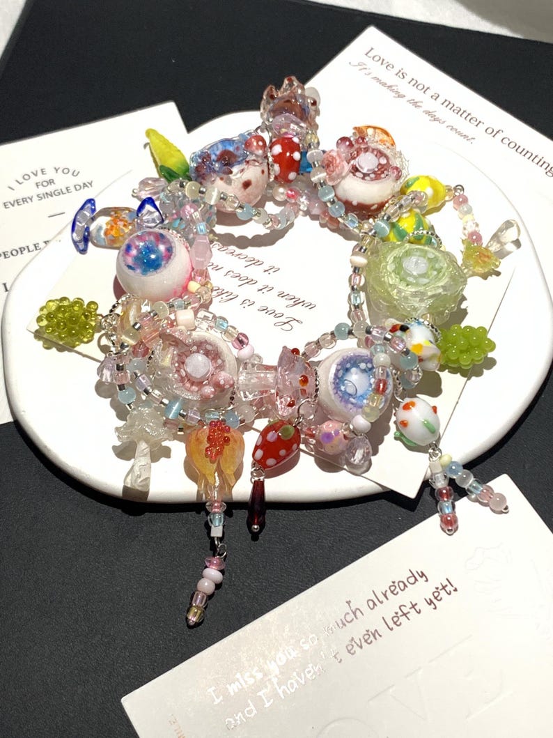 May include: A colorful beaded bracelet with various glass charms, including eye-shaped beads, mushroom-shaped beads, and floral accents. The bracelet is displayed on a white plate with text that reads "I LOVE YOU FOR EVERY SINGLE DAY" and "Love is not a matter of counting."