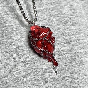 May include: A red, heart-shaped pendant necklace with a silver chain. The pendant is encased in a silver chain and has a dripping effect. The necklace is displayed against a gray background.