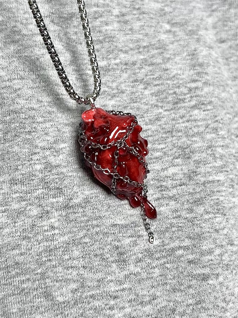 May include: A red, heart-shaped pendant necklace with a silver chain. The pendant is encased in a silver chain and has a dripping effect. The necklace is displayed against a gray background.