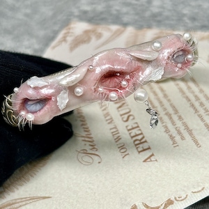 May include: A hair clip featuring a pale, flesh-toned, textured surface with two eye-like openings, each surrounded by fine eyelashes. The piece is adorned with small white pearls and a dangling clear crystal charm, set against a printed paper background.