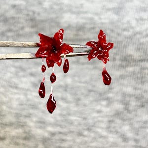 May include: A pair of deep red, glossy flower-shaped earrings. The larger flower features three dangling, teardrop-shaped charms suspended by thin filaments, while the smaller flower has a single teardrop charm hanging from its center.