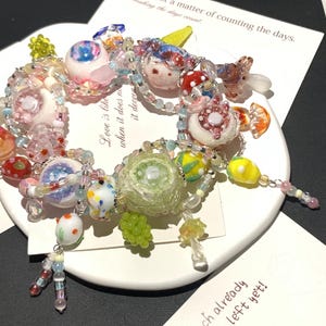 May include: A colorful beaded bracelet with various glass charms, including mushroom, fruit, and floral designs. The bracelet features a mix of pink, white, yellow, and green beads. The bracelet is on a white plate with text that reads "Love is not a matter of counting the days."