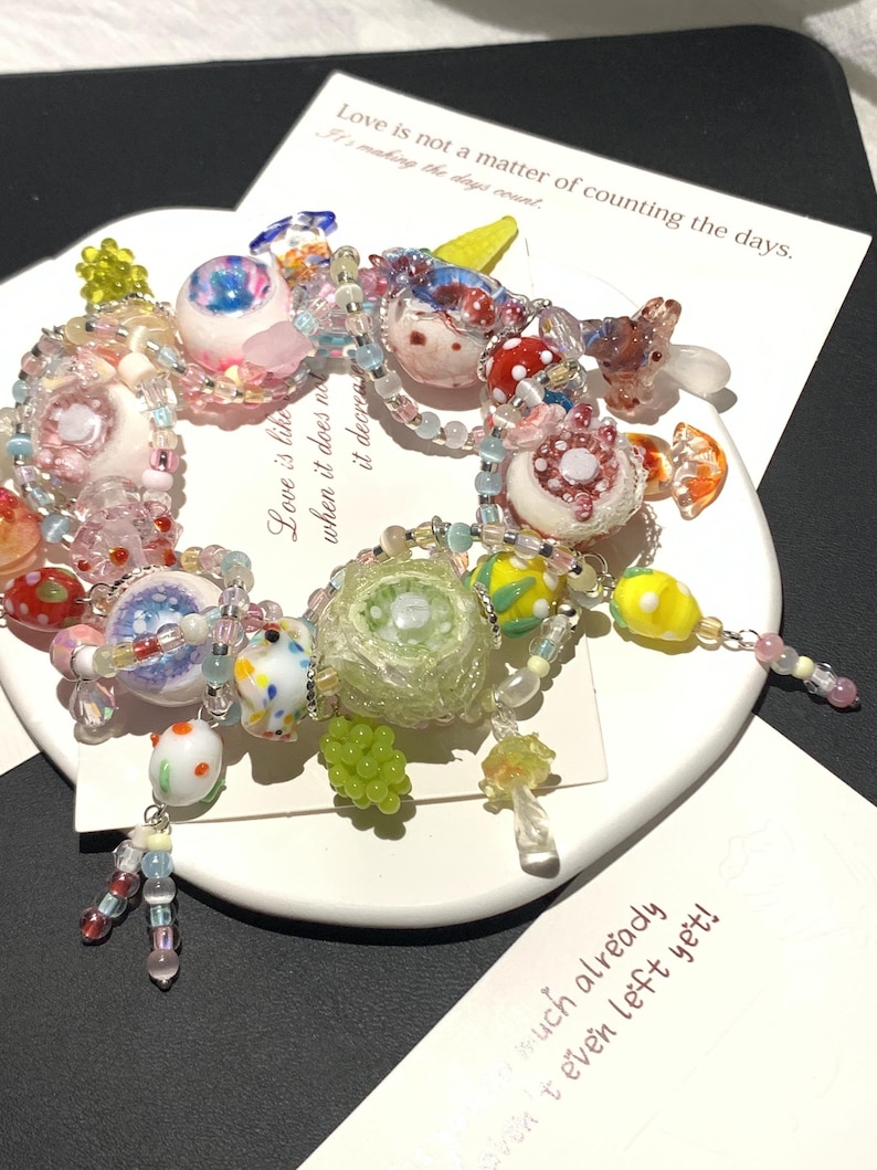 May include: A colorful beaded bracelet with various glass charms, including mushroom, fruit, and floral designs. The bracelet features a mix of pink, white, yellow, and green beads. The bracelet is on a white plate with text that reads "Love is not a matter of counting the days."