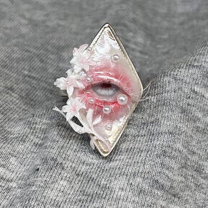 May include: A diamond-shaped ring featuring a central realistic eye illustration with pink shading, surrounded by small white floral accents and tiny white pearls embedded on a glossy, light-colored surface with a metallic silver border.
