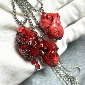 May include: Three necklaces with pendants shaped like realistic red hearts. The pendants are detailed with veins and textures, and are suspended from silver-colored chain necklaces. One heart is wrapped in a silver chain.