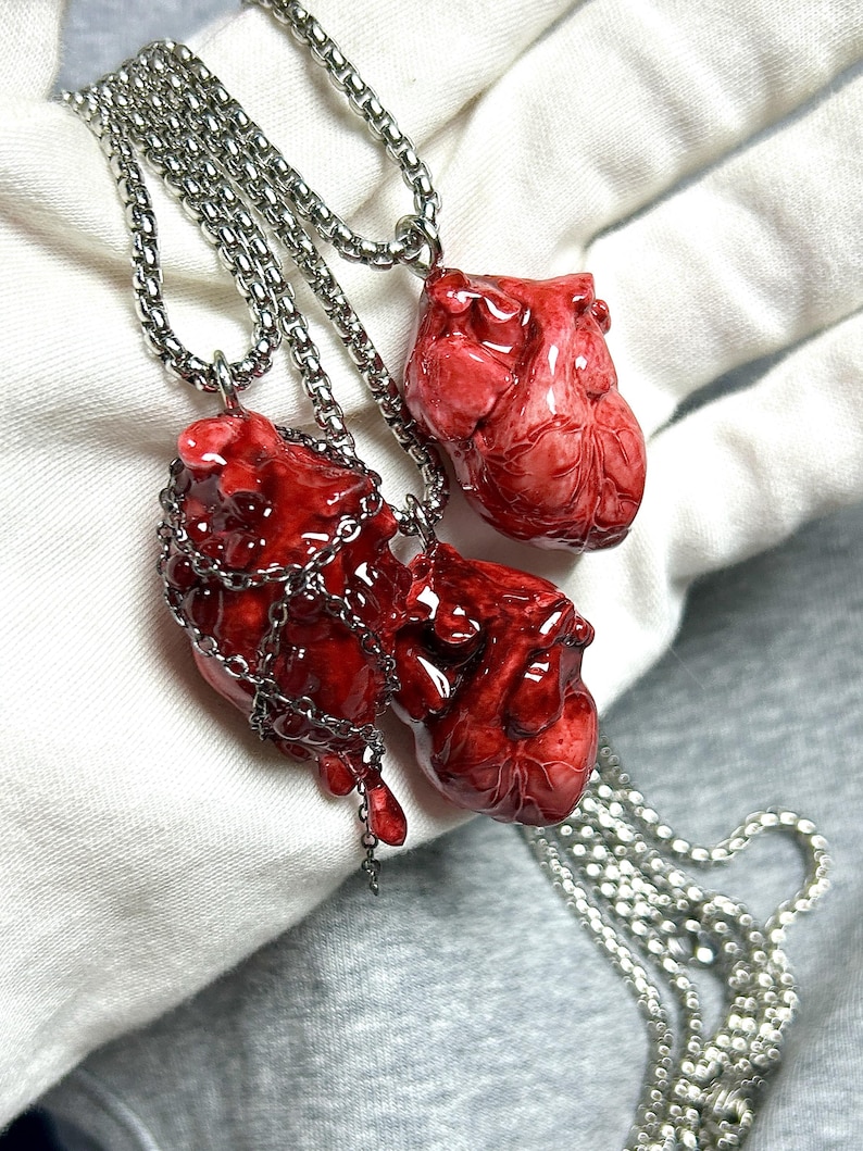 May include: Three necklaces with pendants shaped like realistic red hearts. The pendants are detailed with veins and textures, and are suspended from silver-colored chain necklaces. One heart is wrapped in a silver chain.