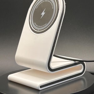 Peut inclure: Un support de charge sans fil blanc avec une bordure noire et un socle de charge circulaire. Le socle comporte un symbole d'éclair et du texte. Le support est conçu pour maintenir un téléphone en charge. Le support est en plastique.