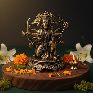 Puede incluir: Una estatua de bronce de una deidad con múltiples rostros y brazos, sosteniendo varios objetos. La estatua está sobre una plataforma de madera, rodeada de flores naranjas, lirios blancos y una pequeña vela encendida. El fondo es oscuro.
