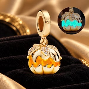 Può includere: Un ciondolo a forma di zucca color oro con una contromaglia decorata da un piccolo cuore e foglie con pietre trasparenti. Il corpo della zucca presenta sezioni in smalto arancione e una funzione luminescente, mostrata in un cerchio color ciano.