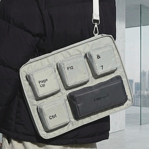 May include: A light grey messenger bag with several square pockets designed to look like keyboard keys. The pockets are labelled with text including "Page Up", "F12", "Ctrl", "& 7", and "Enter". It has a white shoulder strap.