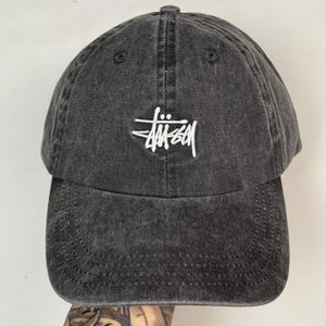 May include: A dark grey baseball cap with a curved peak. The cap has a white embroidered logo on the front. The hat is made of a soft, worn material and has a classic six-panel design. A casual accessory for everyday wear.