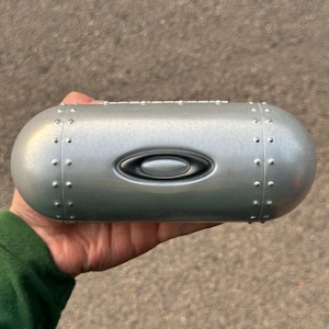 May include: A silver-colored, oval-shaped eyewear case with a prominent Oakley logo. The case has a metallic finish with small, evenly spaced rivets along the edges. The case is held in a person's hand.